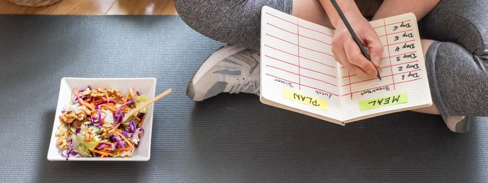 Woman sitting cross legged on a yoga mat next to her salad, while writing in her weekly mean plan.